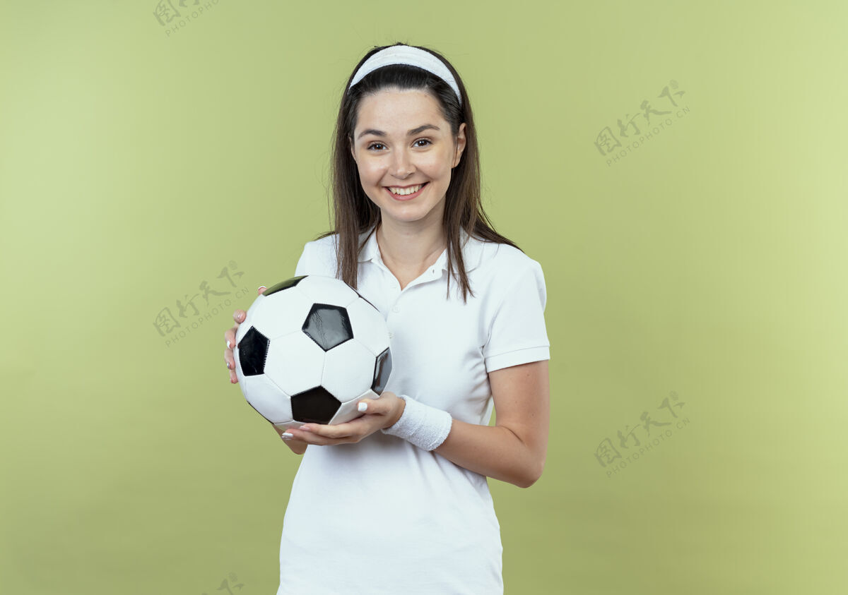 轻戴着头巾 手持足球 面带微笑 积极向上的年轻健身女士站在轻盈的墙上年轻头带持有