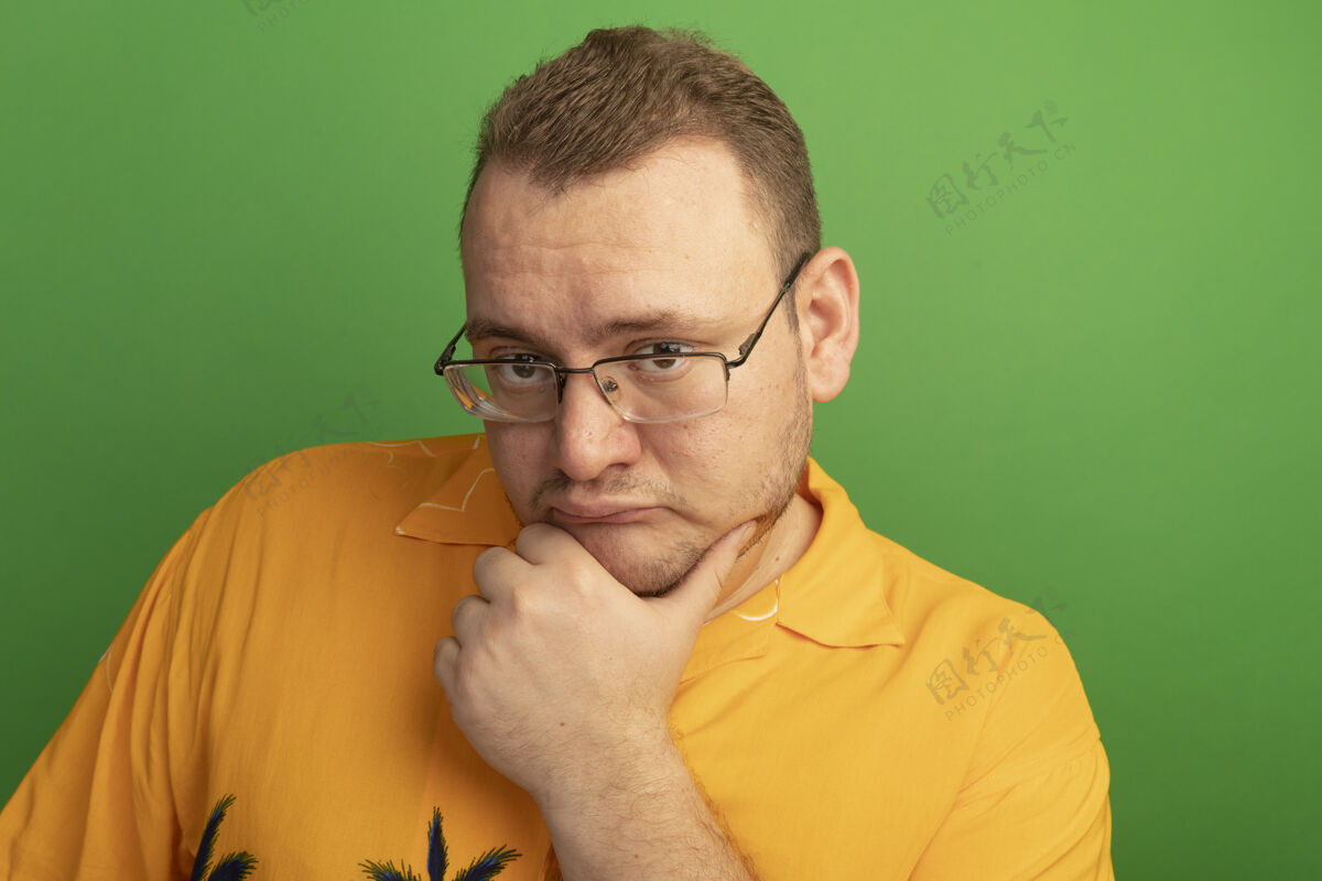 沉思一个戴着眼镜 穿着橙色衬衫 脸上带着沉思的表情的男人站在绿色的墙上思考着眼镜脸男人