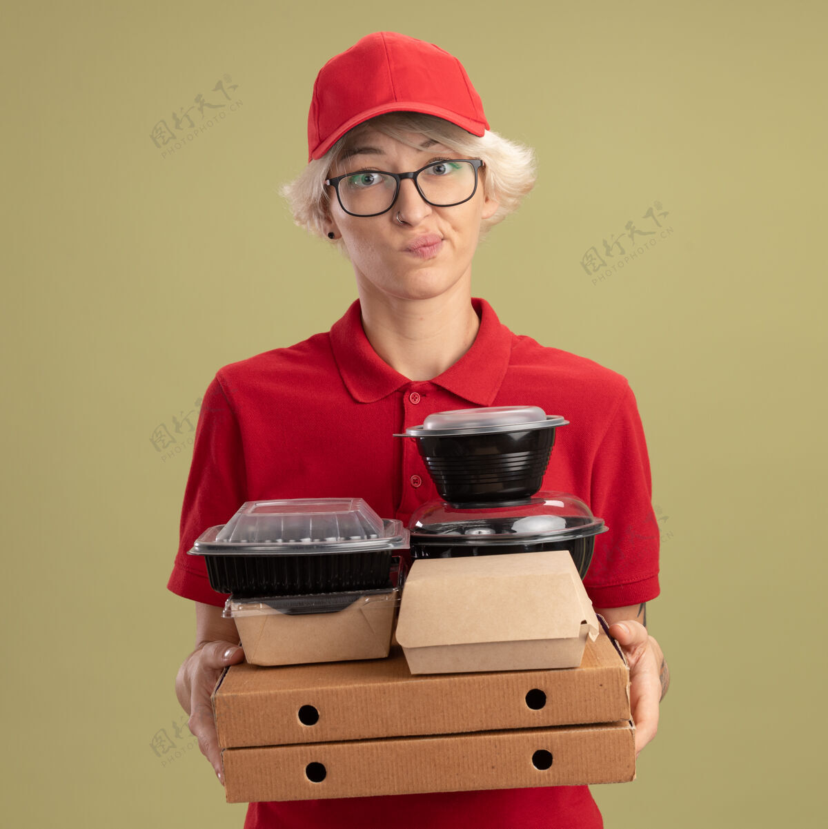 女人年轻的送货员身穿红色制服 戴着帽子 戴着眼镜 手里拿着披萨盒和食品包 满脸怀疑的表情 噘着嘴唇站在绿色的墙上穿送货盒子