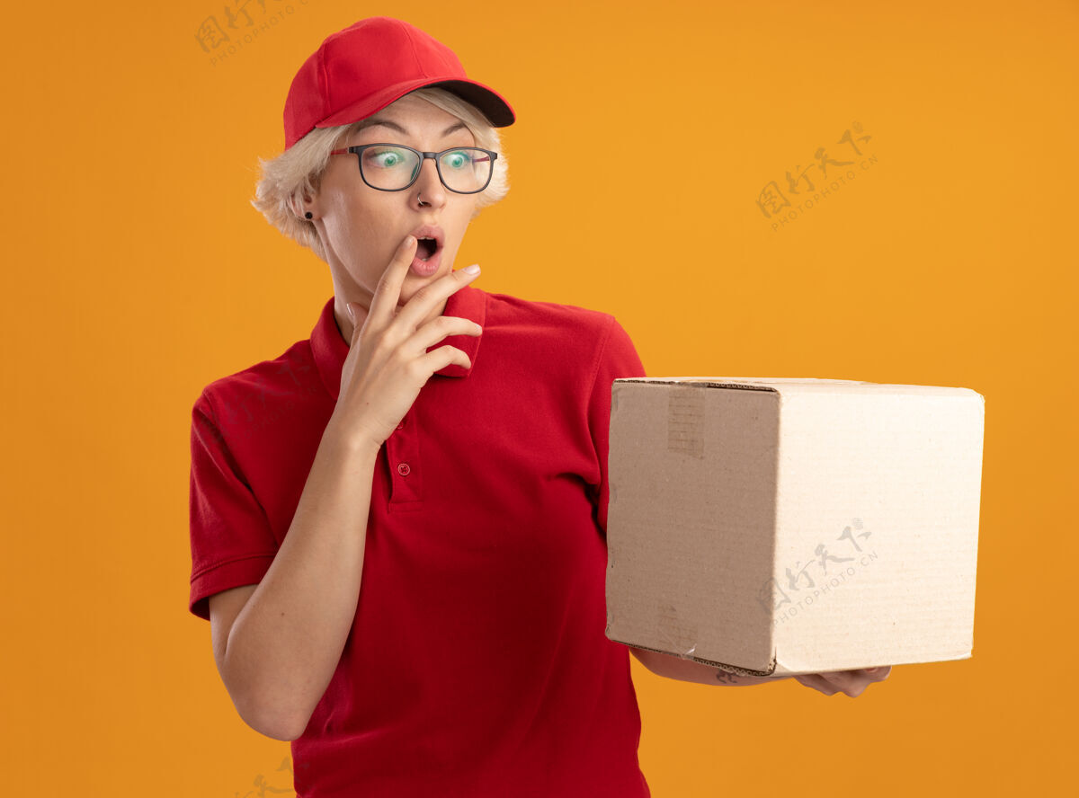 穿上年轻的送货员身穿红色制服 戴着帽子 戴着眼镜 手里拿着纸箱 站在橘色的墙上惊讶地看着它眼镜送货女人