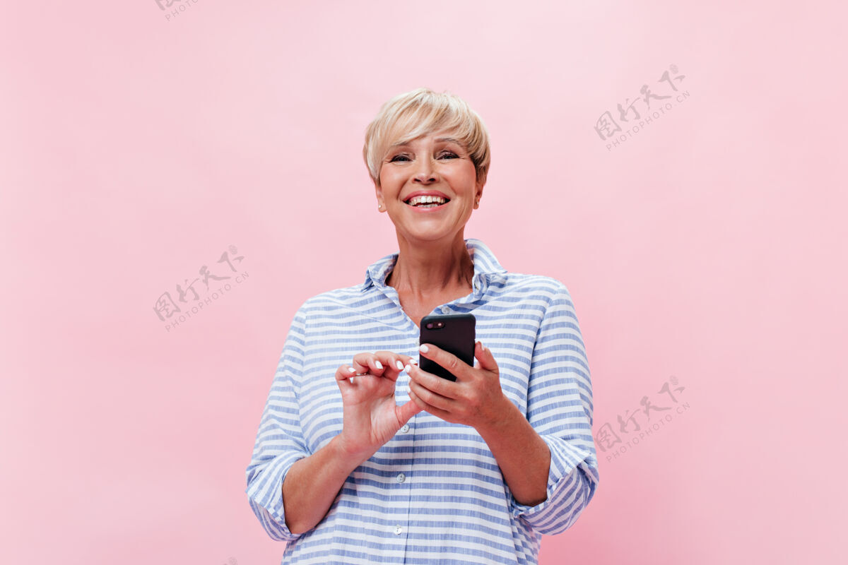 成人身着格子衬衫的美女在粉色背景上愉快地摆出姿势 手里拿着智能手机年龄漂亮护理