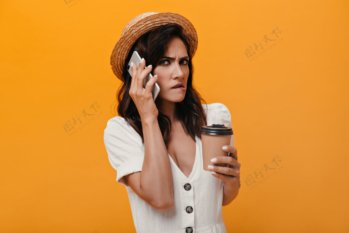圆点不高兴的女孩在橙色背景上讲电话 手里拿着一杯咖啡脸衬衫苍白