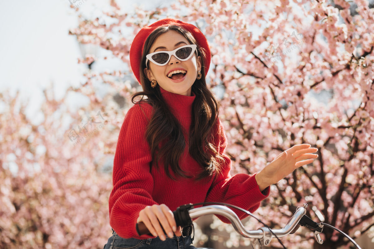 肖像在樱花的背景下 美丽的女孩穿着红色的衣服 戴着太阳镜快乐的女人穿着羊绒衫 戴着贝雷帽 微笑着骑着自行车美女天女孩