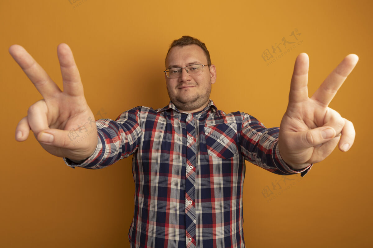 检查一个戴着眼镜 穿着格子衬衫的男人兴高采烈地笑着 橙色的墙上挂着v字标志眼镜欢呼手势