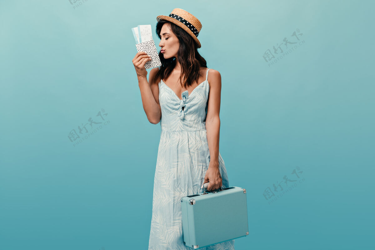手提箱戴草帽的漂亮女士拿着蓝色背景下的时髦包和票穿着夏装摆姿势的漂亮女人肖像思考女孩