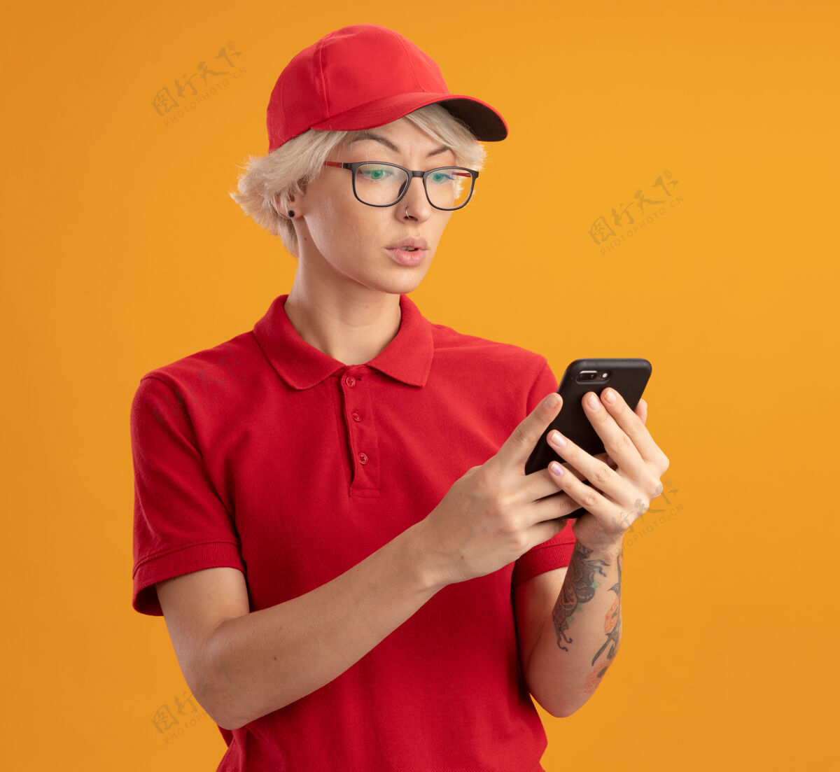 制服身穿红色制服 戴着眼镜 面带严肃表情的年轻送货员站在橙色的墙上看着自己的智能手机帽子穿女人