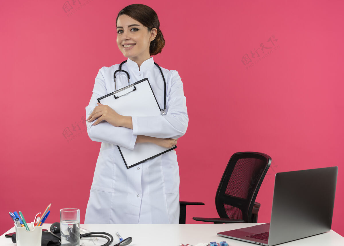 年轻面带微笑的年轻女医生穿着医用长袍 听诊器站在办公桌后面 带着医疗工具和笔记本电脑 把剪贴板隔离在粉红色的墙上剪贴板笔记本电脑女