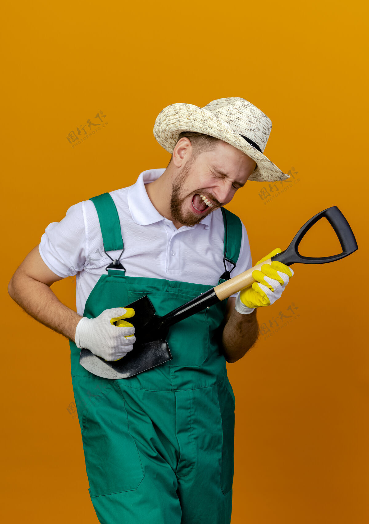 玩年轻英俊的斯拉夫花匠 穿着制服 戴着帽子 戴着园艺手套 手里拿着黑桃 闭着眼睛 假装在橙色的墙上弹吉他手套持有年轻