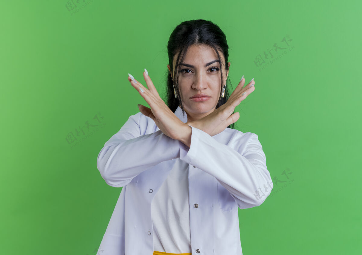穿着严格的年轻女医生穿着医学长袍做没有手势隔离在绿色的墙壁与复制空间女性严格医生