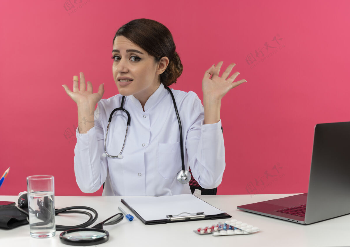 办公桌穿着医用长袍和听诊器的年轻女医生坐在桌子旁 手里拿着医疗工具和笔记本电脑 两手空空的 被隔离在粉红色的墙上穿听诊器坐着