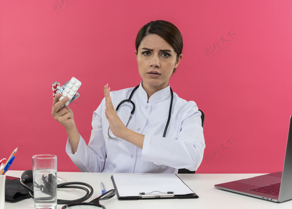 坐着皱眉的年轻女医生穿着医用长袍和听诊器坐在办公桌旁 手里拿着医疗工具和手提电脑 手里拿着药品 并在粉色的墙上做着不孤立的手势听诊器医生皱眉