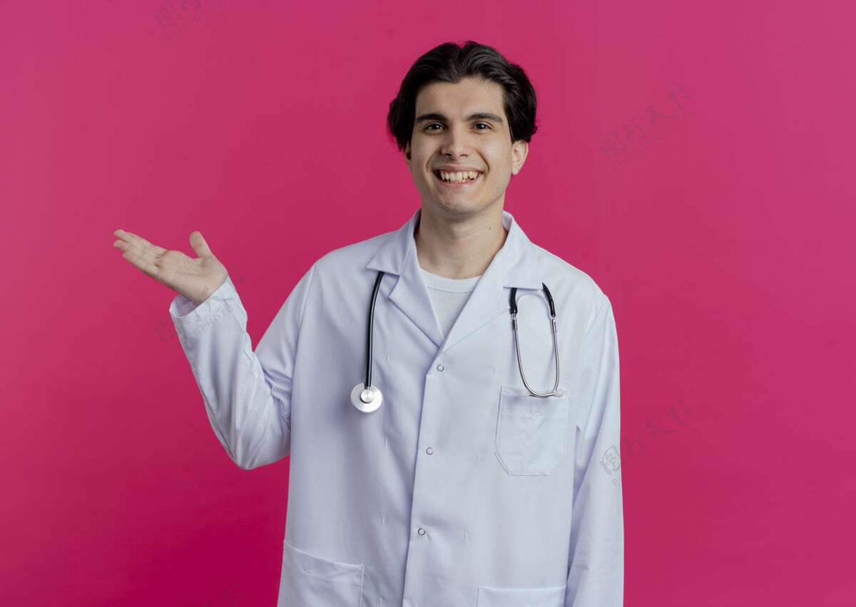 医学微笑的年轻男医生穿着医用长袍和听诊器 在粉红色的墙上显示出一只空手 并留有复印空间手长袍穿