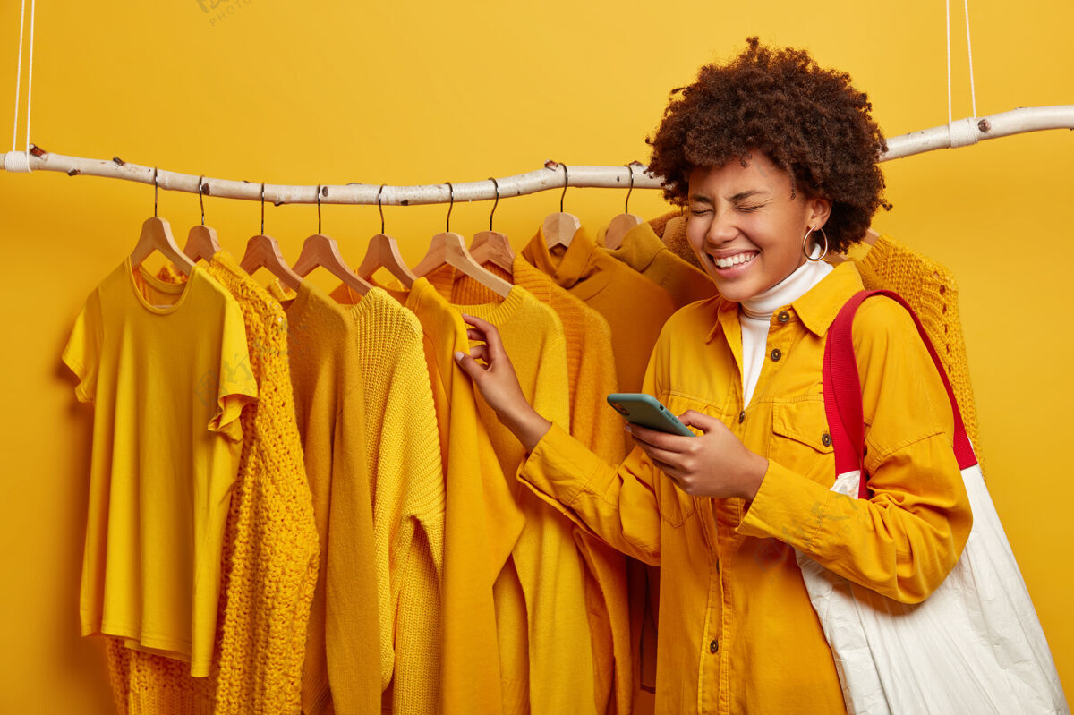 年轻人非洲女人穿着时髦的黄色夹克 拎着购物袋 用手机上网交流 在黄色背景下的服装栏杆旁摆姿势应用程序装装自拍