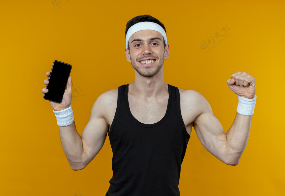 运动戴着头巾的年轻运动型男子展示智能手机握紧拳头 为橙色感到高兴和兴奋握紧显示头带