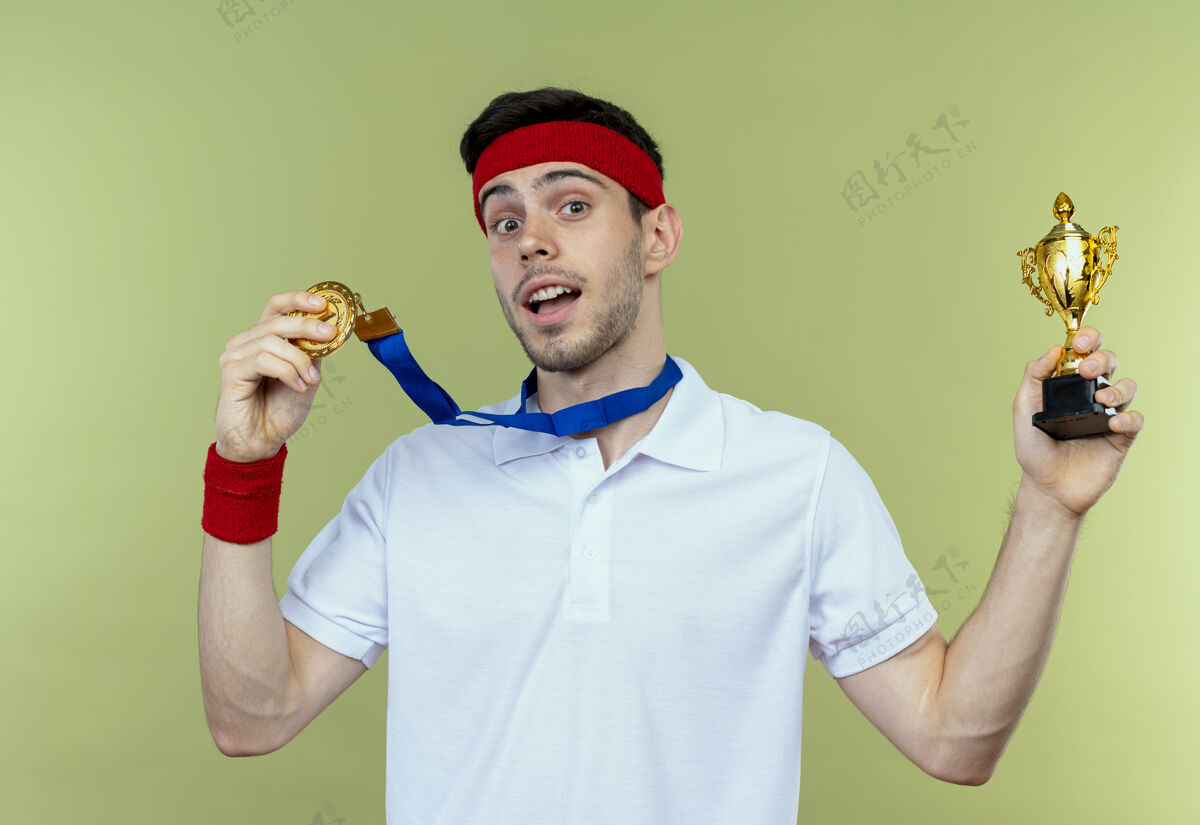 周围戴着头巾 脖子上戴着金牌的年轻运动型男子手持奖杯 为绿色而高兴和兴奋兴奋奖杯奖牌