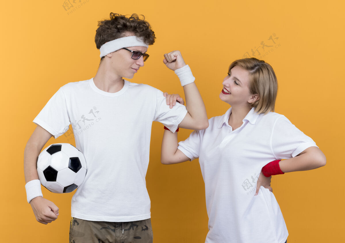 站着一对年轻的运动型情侣展示着他拿着足球的肱二头肌和他微笑的女友在橘子上橙色表演足球