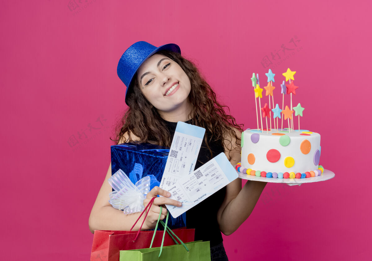 抱着一位年轻漂亮的女士 卷发 戴着节日礼帽 手里拿着生日蛋糕礼盒和机票 开心地笑着 生日派对的概念超过粉色聚会头发礼物