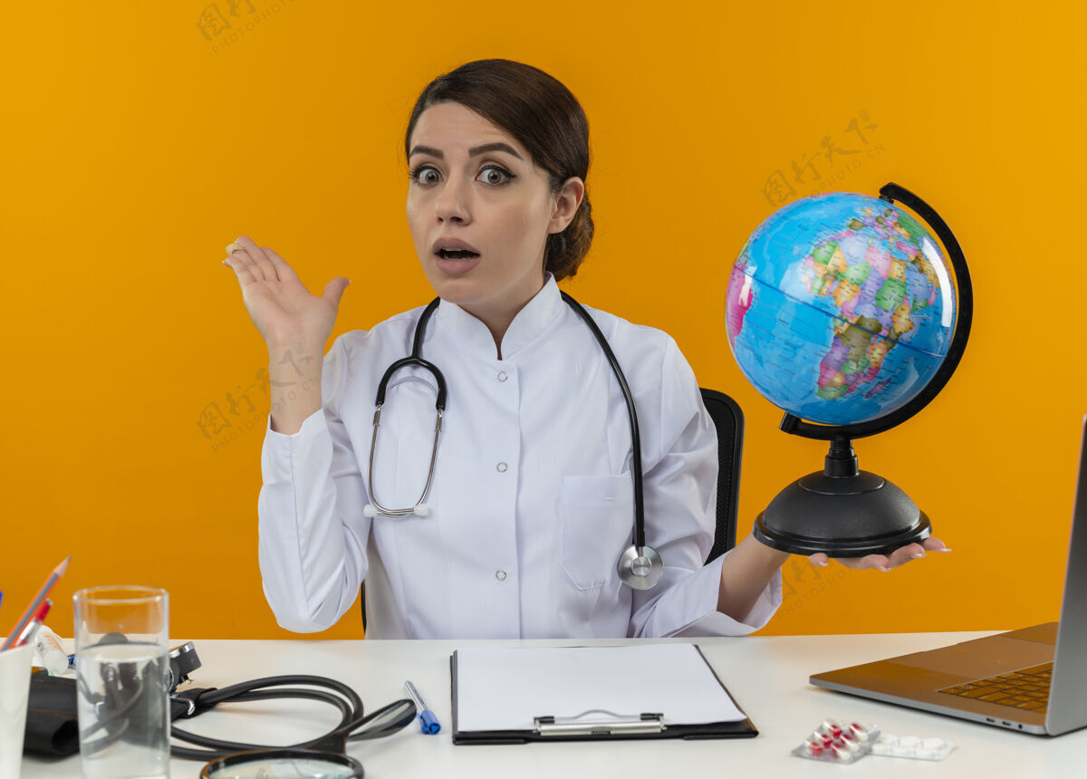 医疗年轻的女医生穿着医用长袍 手持听诊器 坐在桌子旁 手里拿着医疗工具和笔记本电脑 手里拿着地球仪 两手空空健康女性年轻