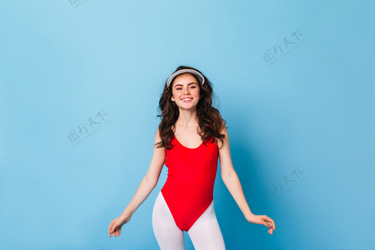 80后风格红润脸蛋的年轻女子穿着红色紧身衣和运动紧身裤 面带微笑地看着前面的蓝色墙壁模特帽子化妆