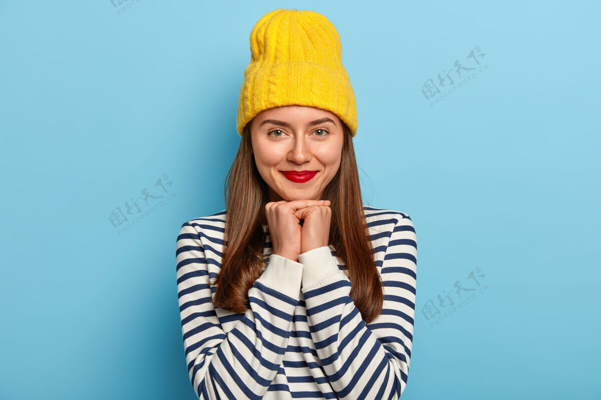 人温柔好看的黑发女人站在蓝色背景的黄色帽子和条纹毛衣 穿红色唇膏姿势乐观毛衣