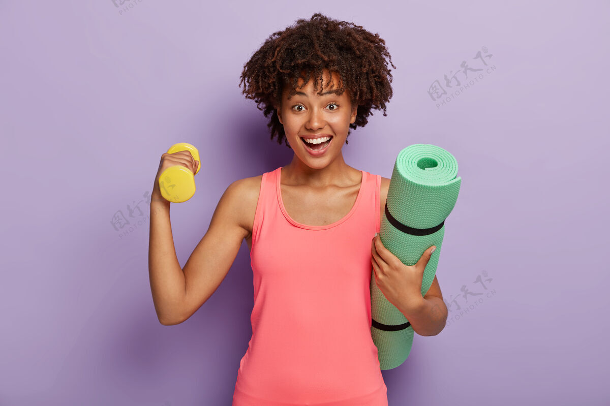 卷发头发清爽的快乐女人 举哑铃训练肌肉 穿着粉色上衣 背着绿色的健身垫私人教练活跃哑铃