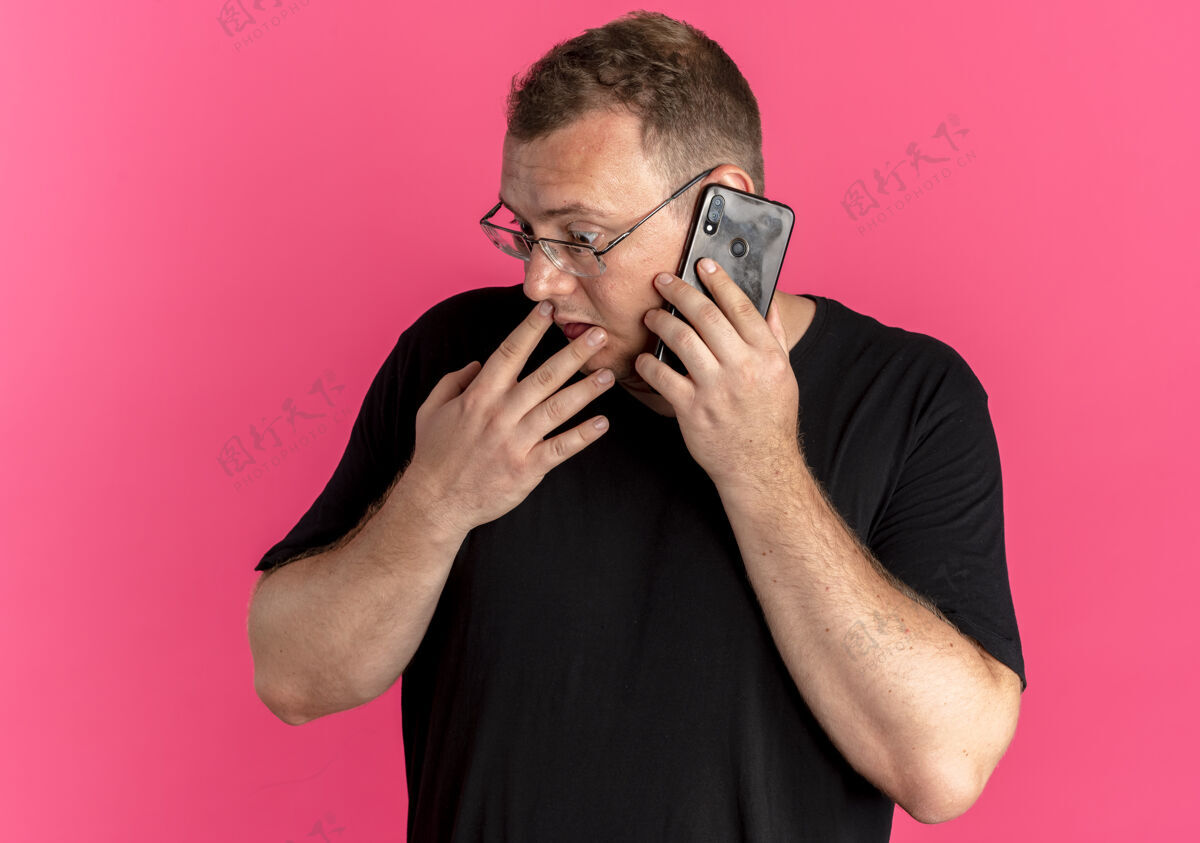 边戴眼镜的超重男子身穿黑色t恤 一边困惑地看着一旁 一边用粉色手机交谈男士边电话