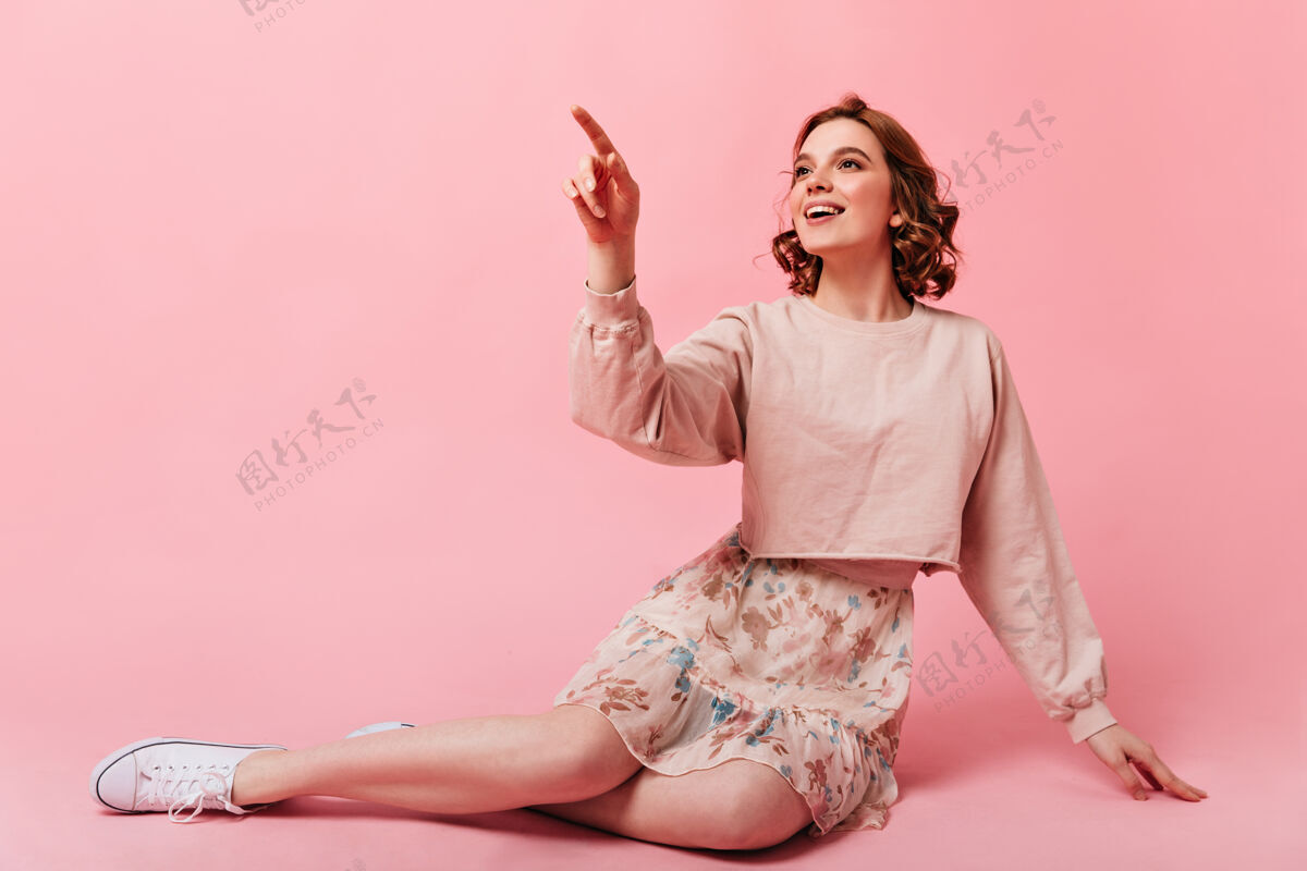 优雅迷人的女孩穿着裙子坐在粉红色的背景上微笑着灵感来源于年轻的卷发女子用手指着休闲一个人女性