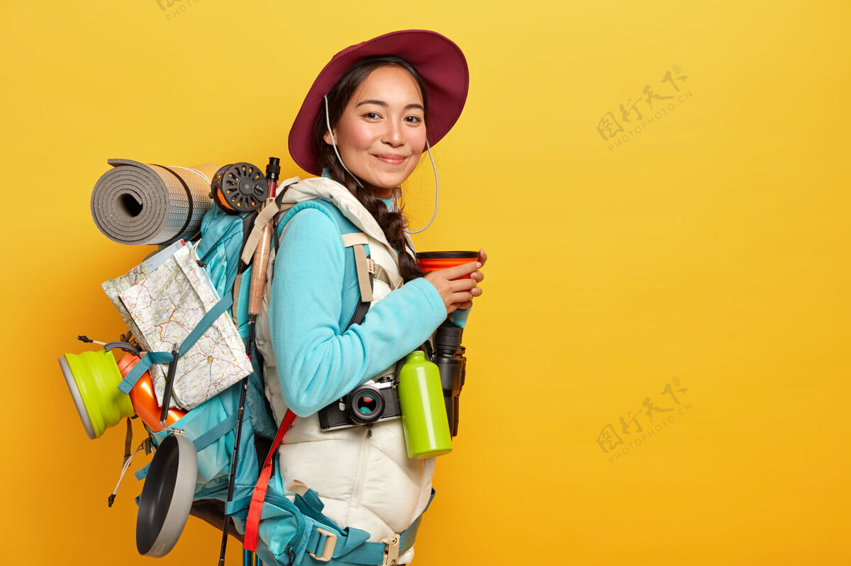 饮用快乐的年轻亚洲女游客在路上停下来喝咖啡 戴帽子 穿休闲装 背着背包 长途旅行 探索新的地方 喜欢旅游瓶子看背包