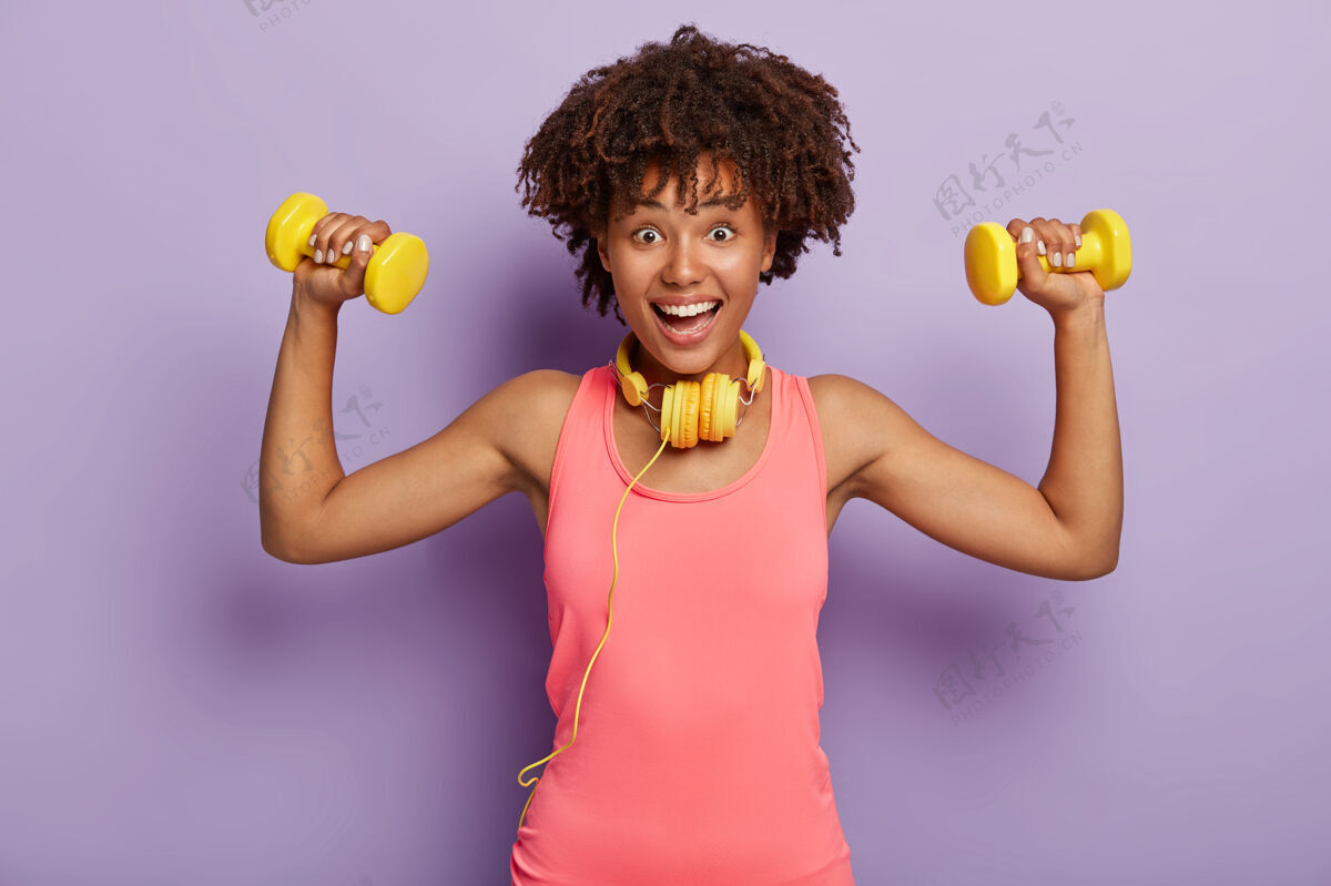 能量积极的女人留着非洲式发型 举着哑铃 戴着黄色耳机和粉色背心 在紫色的工作室墙上摆姿势卷发运动女人有氧运动