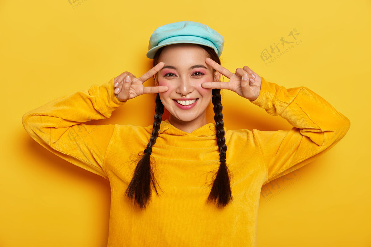 化妆无忧无虑微笑的亚洲女士在眼睛附近做出胜利和平的手势 心情愉快 微笑温柔 化妆生动 戴着时髦的帽子和运动衫 隔离在黄色的墙上女性时尚运动衫