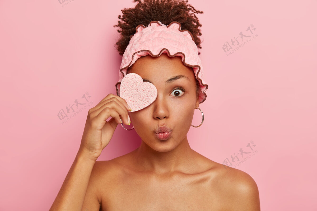 水疗惊讶的非洲女人用化妆海绵遮住一只眼睛 保持嘴唇圆润 眼睛被窃听 在spa沙龙做美容 梳卷发自然服装惊讶