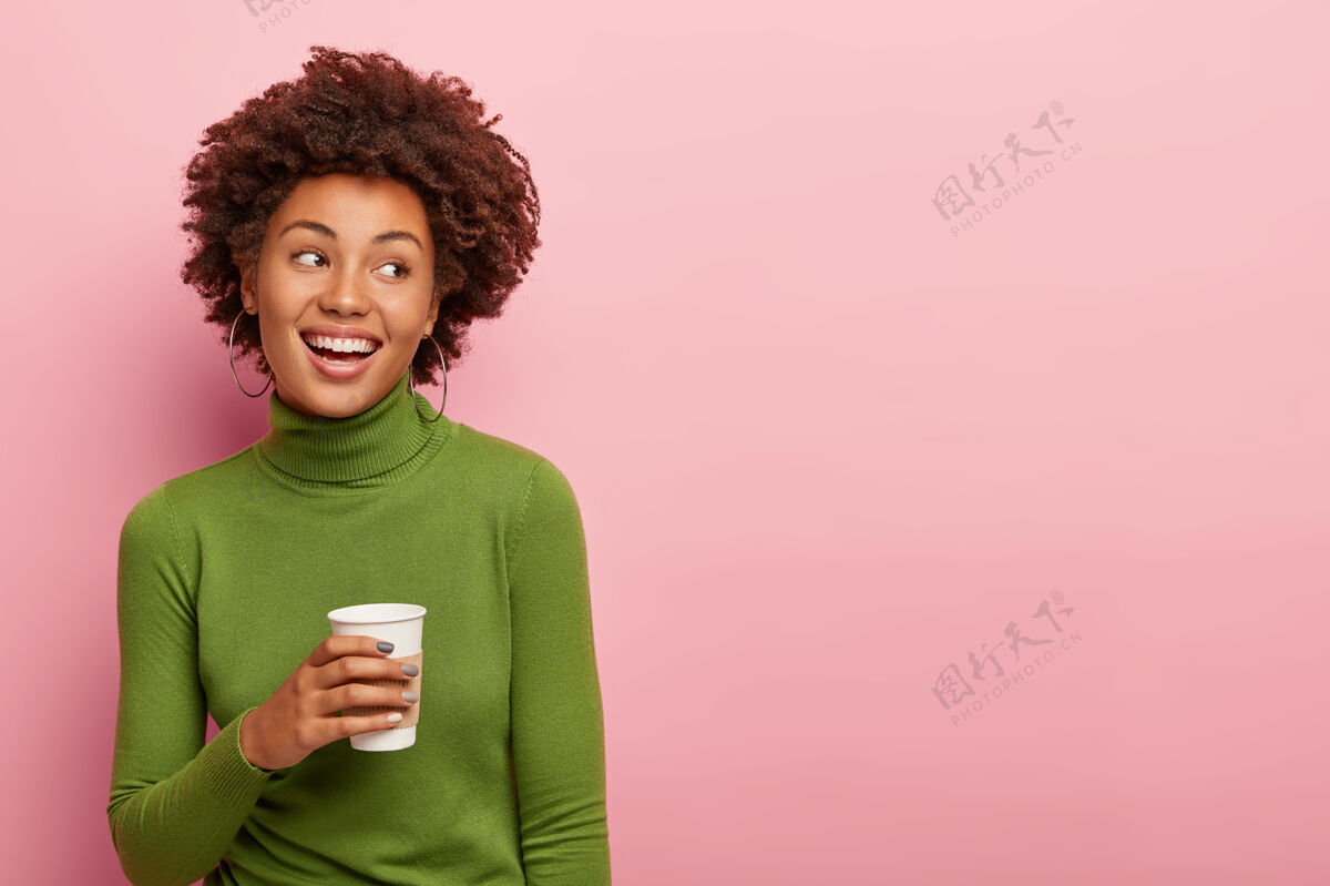非洲可爱的卷发女人拿着外卖咖啡 享受休息 穿着绿色圆领毛衣 看着右边 对着粉色的墙壁摆姿势 为你的广告内容腾出空间姿势肖像理发
