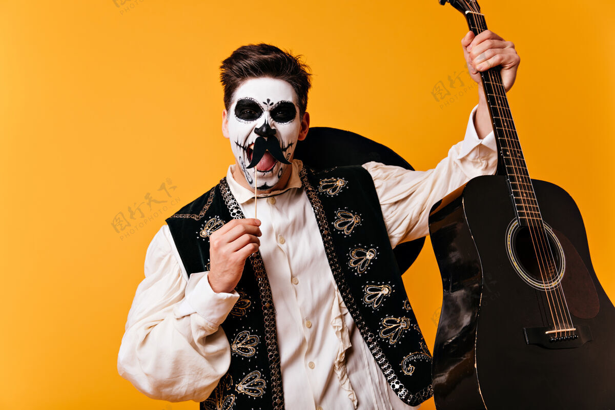 节日墨西哥棕色眼睛的男人 以头骨的形式进行面部艺术 情绪化地大叫 摆出假胡子的姿势 手里拿着吉他 站在橙色的墙上娱乐男人帽子