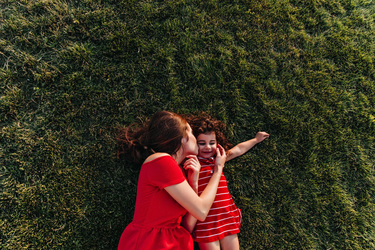 黑发身穿红裙子的黑发女子躺在草地上亲吻着女儿的脸颊户外头顶上年轻母亲和她的孩子在公园里的照片年轻拥抱放松