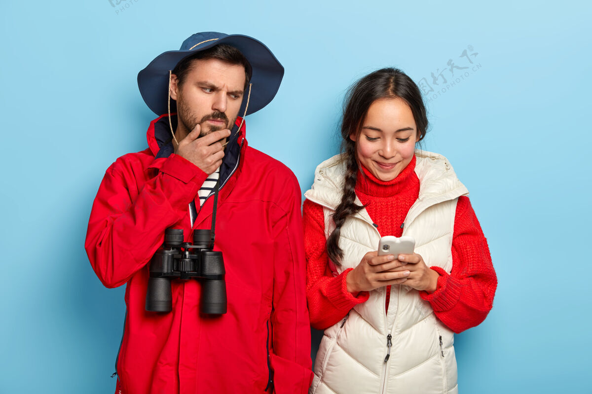 在线不满的男人拉着下巴 愤怒地看着女友的智能手机 穿着休闲服 拿着望远镜和快乐的亚洲女孩打字留言 专注于手机度假家庭女性