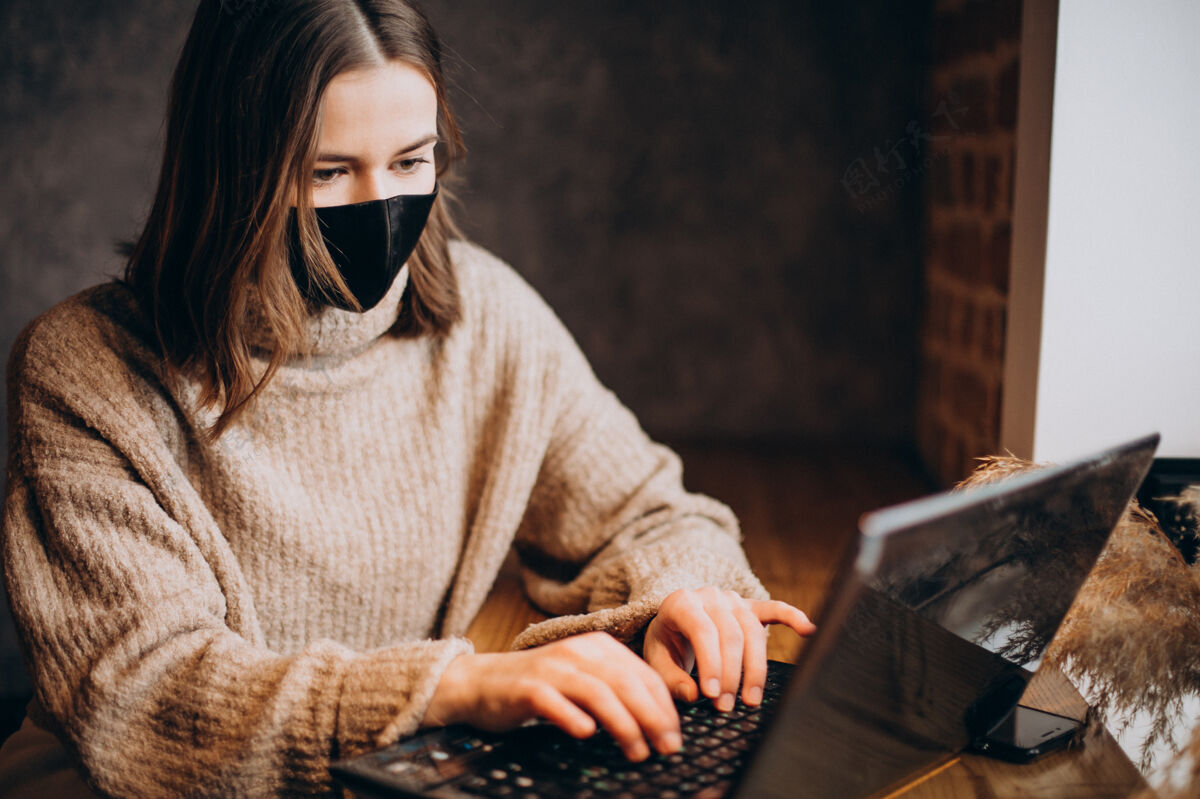 智能戴着面具在咖啡馆里用笔记本电脑工作的年轻女子专业年轻女孩