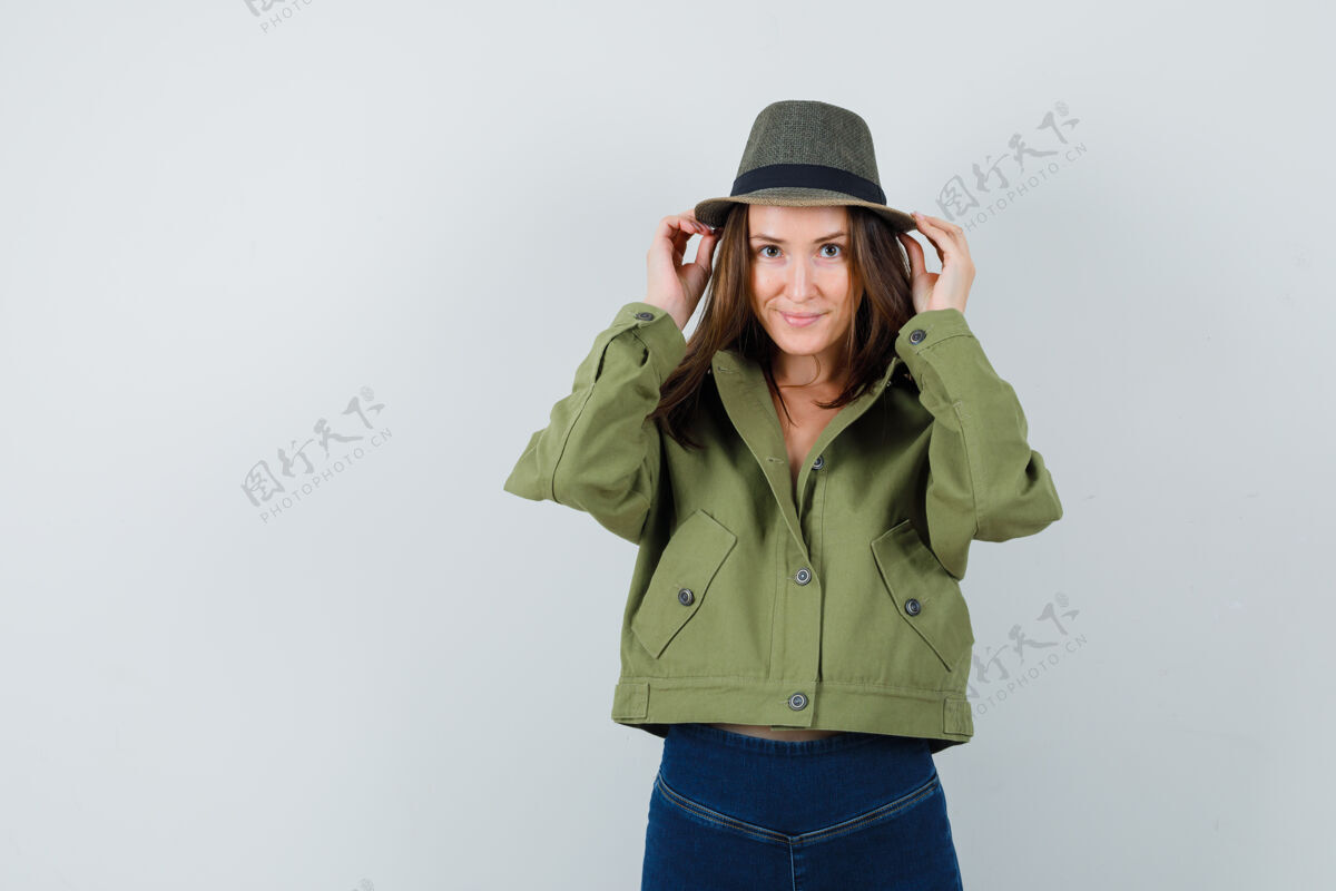 微笑年轻的女性调整她的帽子在夹克 裤子和看起来可爱前视图时尚裤子休闲