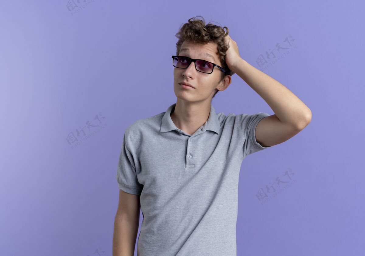 眼镜一个戴着黑眼镜 穿着灰色马球衫 手放在头上的年轻人站在蓝色的墙上看向一边 感到困惑困惑放一边男人
