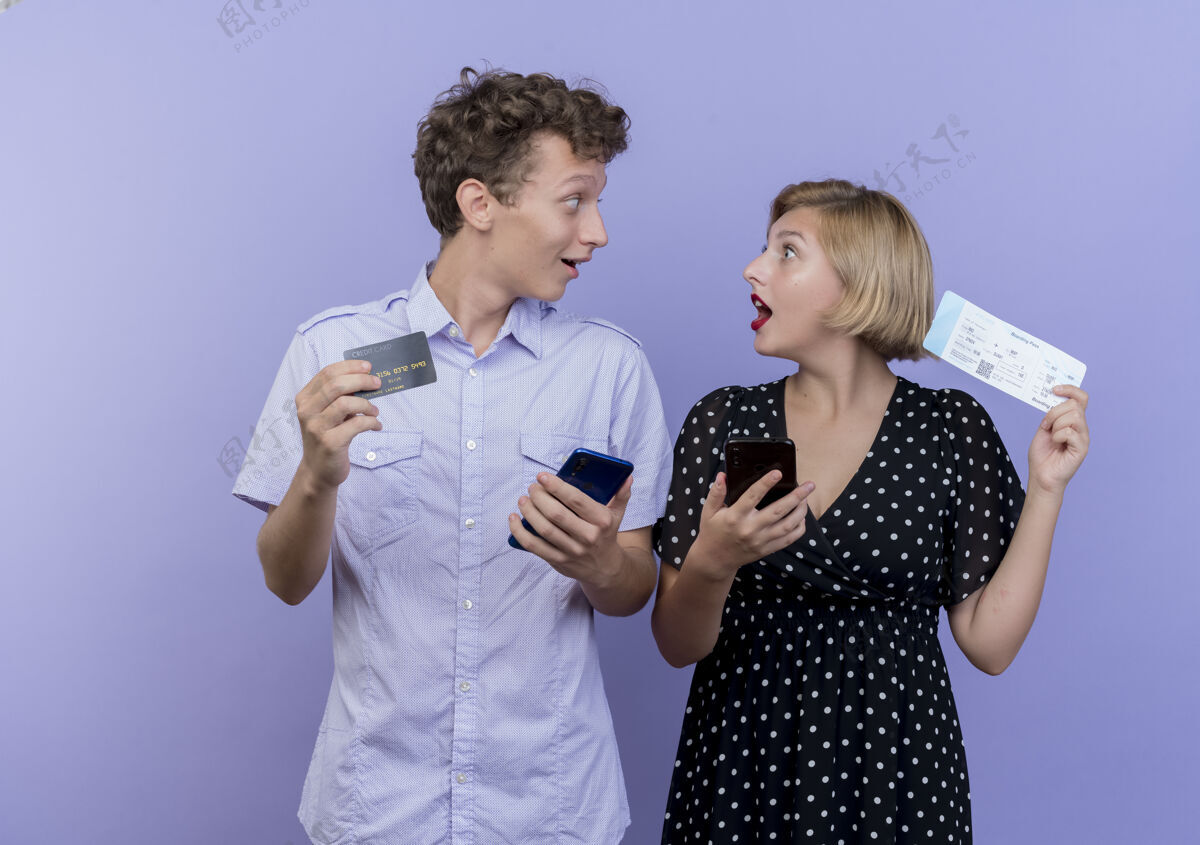期待一对年轻漂亮的夫妇拿着智能手机和信用卡 拿着机票 高兴又惊讶地看着对方越过蓝色的墙壁夫妇每个年轻人