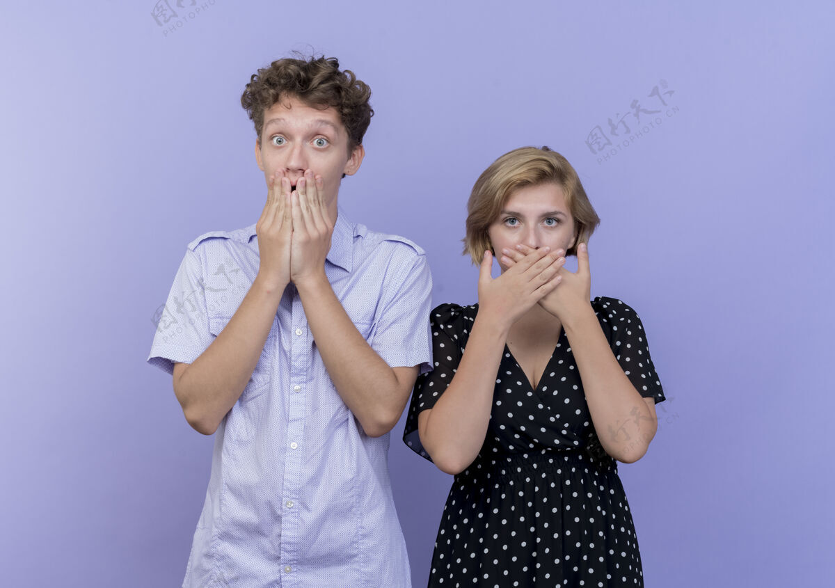 手年轻漂亮的夫妇站在蓝色的墙壁上 双手捂着嘴被惊呆了掩护站年轻人