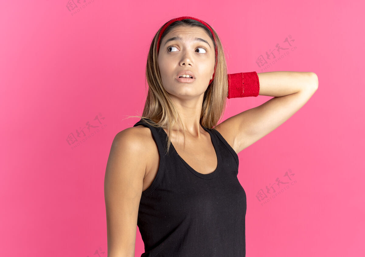 年轻身穿黑色运动服 头戴红色头巾的年轻健身女孩站在粉红色的墙上 茫然地看着一旁健康站旁边