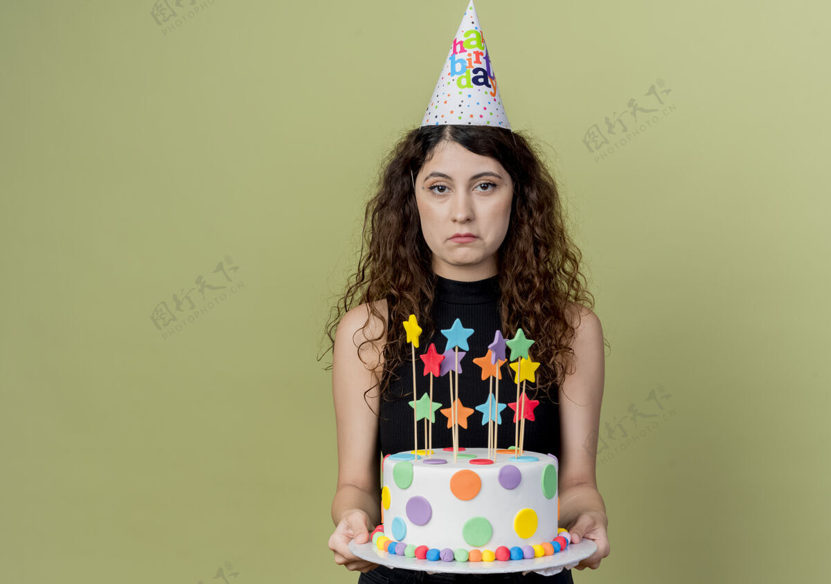 蛋糕戴着节日帽的卷发美女手持生日蛋糕 脸上带着悲伤的表情站在明亮的墙上悲伤女人帽子