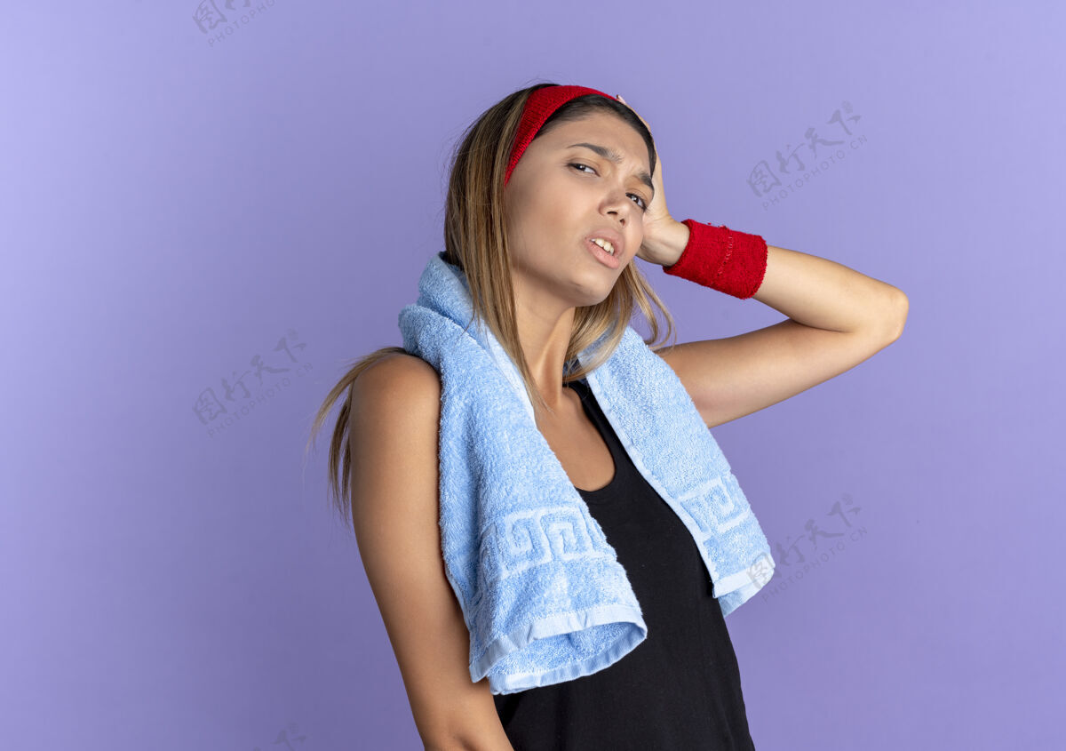 脖子身穿黑色运动服 头戴红色头巾 脖子上缠着毛巾的年轻健身女孩 看上去很困惑 因为她站在蓝色的墙上时 手放在头上 犯了个错误女孩站健身