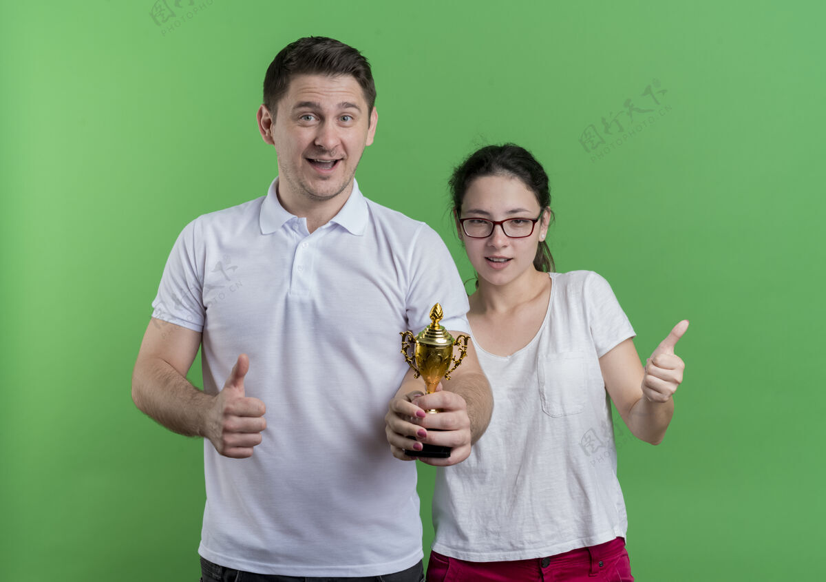 奖杯年轻的运动情侣站在一起举着奖杯竖起大拇指站在绿色的墙上男人表演女人