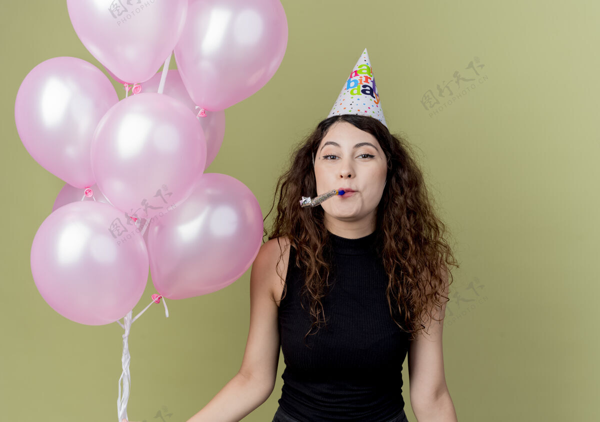 积极一个年轻漂亮的女人 卷发 戴着节日帽 手里拿着气球吹着口哨 快乐而积极地庆祝生日 站在光墙上举行光吹