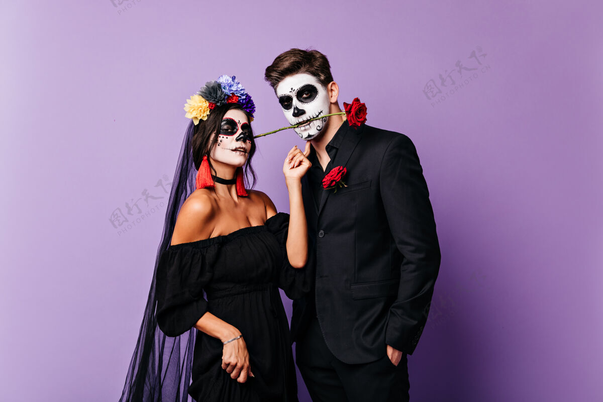 男孩一对身着节日嘉年华服装的情侣在紫色背景上摆姿势的特写照片热情的墨西哥男人用牙齿叼着玫瑰 他的新娘看着镜头浪漫骷髅传统