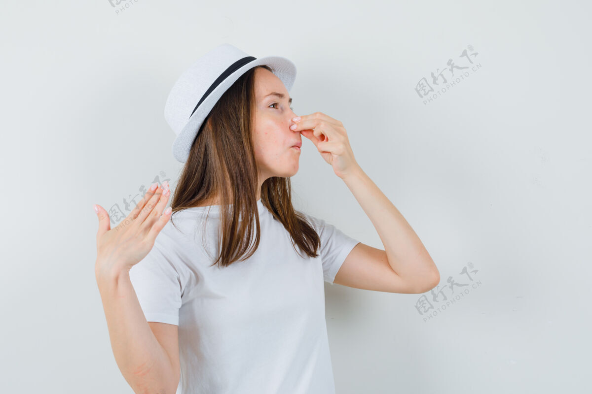 坏年轻女孩穿着白色t恤 戴帽子捏鼻子 由于气味难闻 看起来很恶心 前视捏女人厌恶