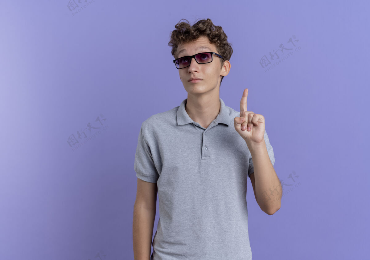 站一个戴着黑眼镜 穿着灰色马球衫的年轻人用食指指着蓝色墙上的东西穿男人目录