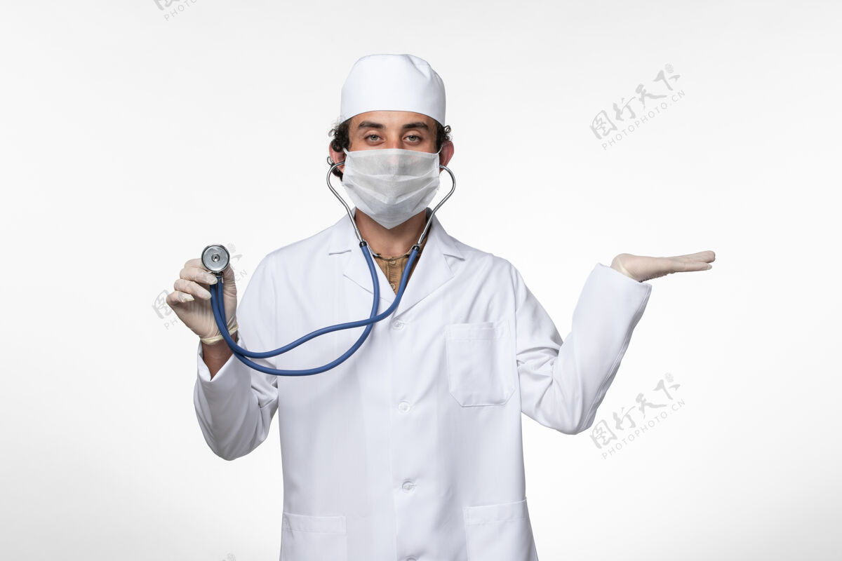 听诊器正面图：男医生 穿着医疗服 戴着口罩 以防感染 用听诊器在浅白色的墙壁上观察疾病 传播病毒男性灯光冠状病毒预防