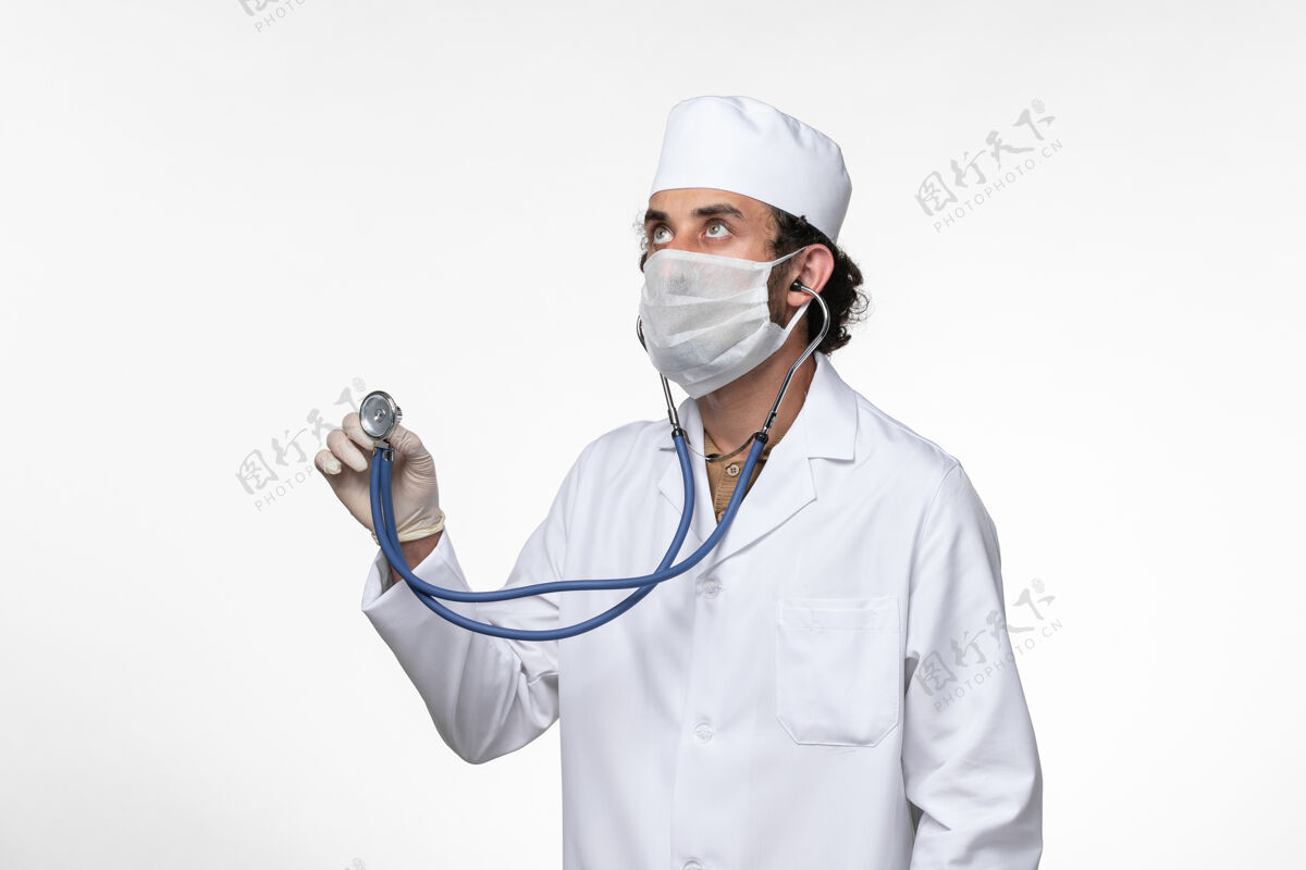 男性正面图男医生穿着医疗服 戴着口罩作为防护罩 用听诊器对白墙病病毒进行了冠状病毒-大流行套装冠状病毒预防使用